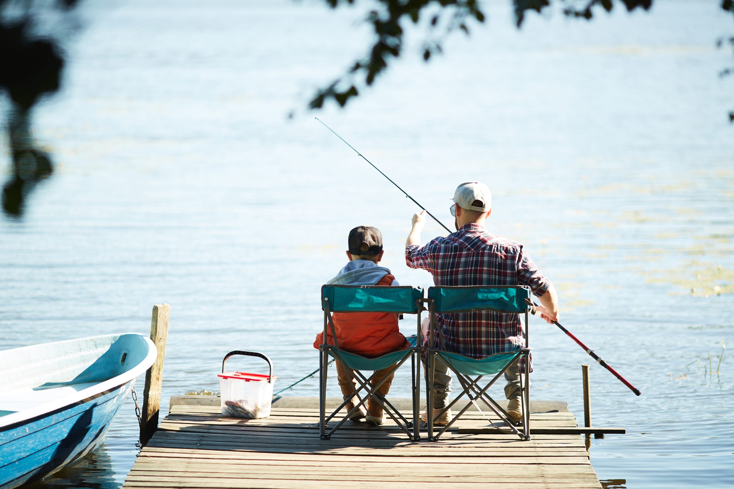 Rear view of man and his son sitting in front of water and fishing together at summer weekend