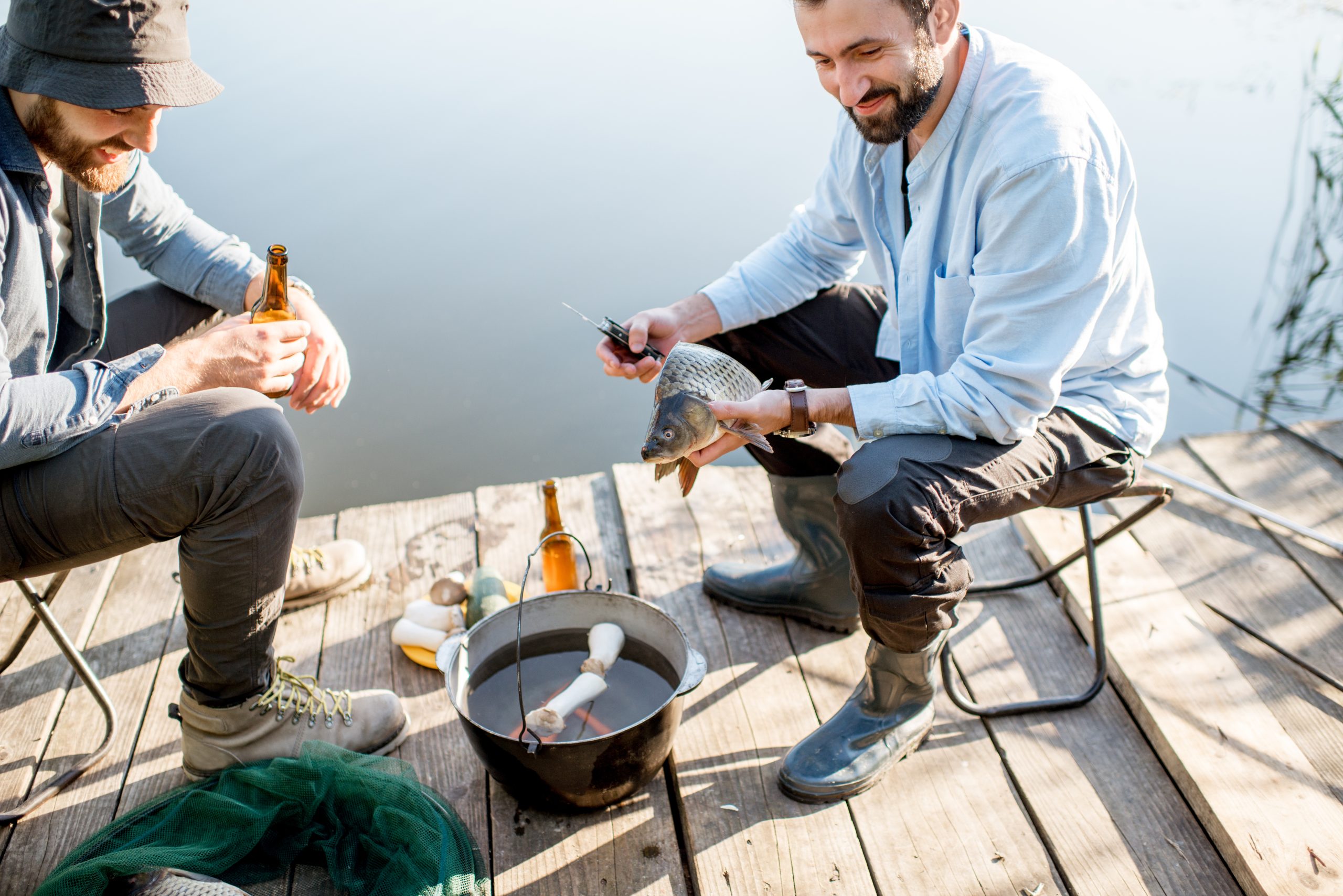 Two friends sitting together with beer and fish on the picnic while fishing near the lake